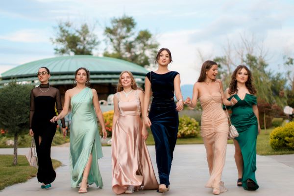 Six young women in long evening gowns walking together while smiling and holding hands