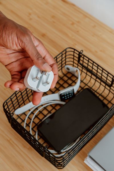 A hand holding a white 3-pin power adapter with a black phone, charging cable, and a smart watch in a basket