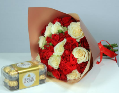 A bouquet of red and white roses wrapped in a dusty pink paper next to a box of chocolates