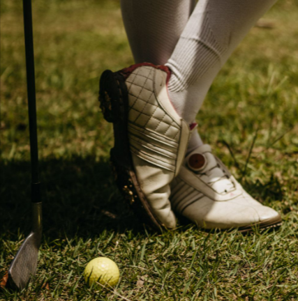 Close-up of golf shoes, club, and ball from Gamola Golf on a green field.