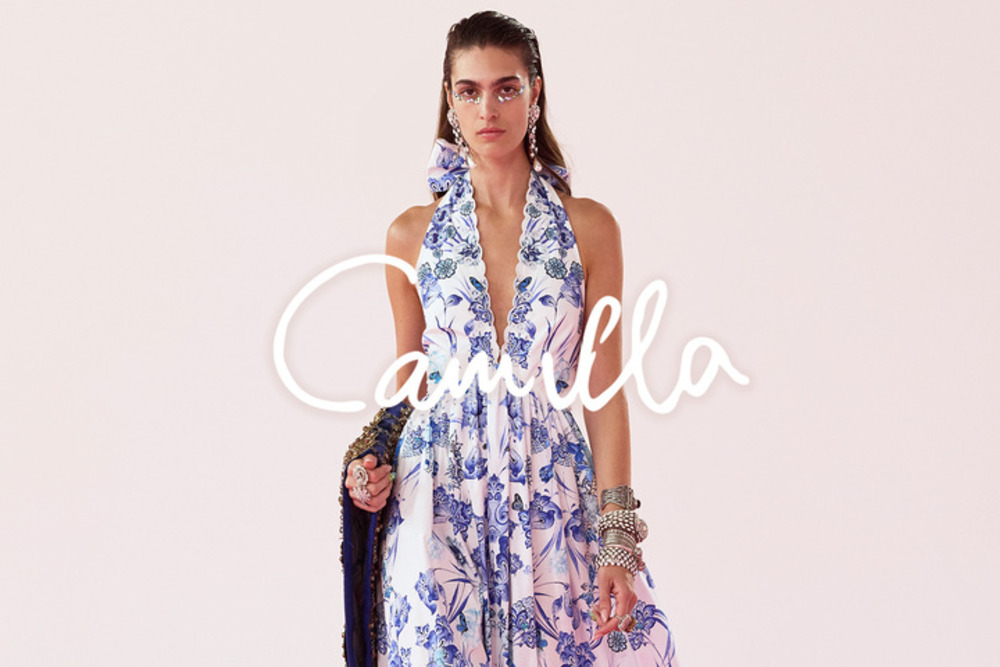 model wearing a blue and white dress from Camilla. Model has decorative makeup and the Camilla name logo can be seen there is also a pink background