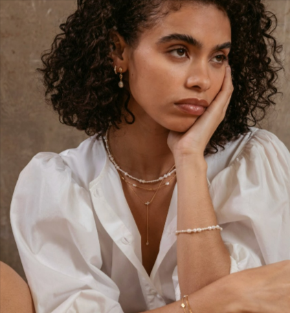 A lady with curly hair wearing a white blouse and wearing various pieces from the Astrid & Miyu jewellery collection