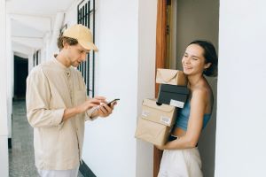 A woman smiling standing by the door holding a stack of parcels while the courier checks his phone