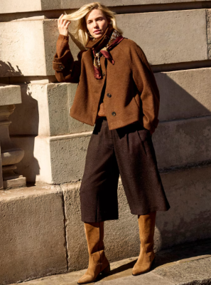 A woman wearing a brown jacket, wide-leg pants and boots standing infront of a stoned wall