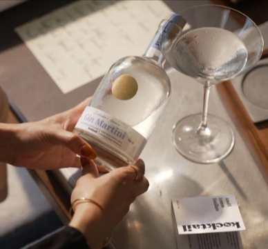 A hand holding a bottle of Kocktail Gin Martini on sale beside a prepared cocktail glass