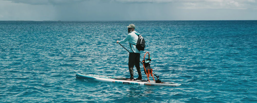 A man paddles while standing on a BOTE paddle board with blue water on the background