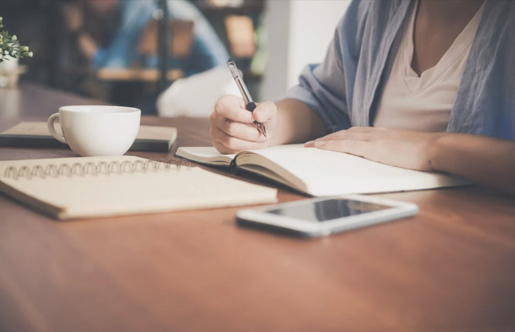 Woman writing in a notebook beside a teacup and tablet computer on a brown desk