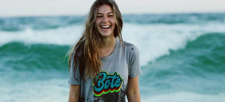 A smiling woman wearing a gray BOTE t-shirts with sea waves in her background