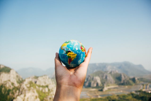 A person holding a tiny plastic world globe while standing in front of a mountain range