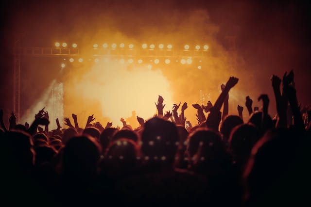 A silhouette of a crowd of people raising their hands at a concert with a cloud of orange smoke and a stage in the background.