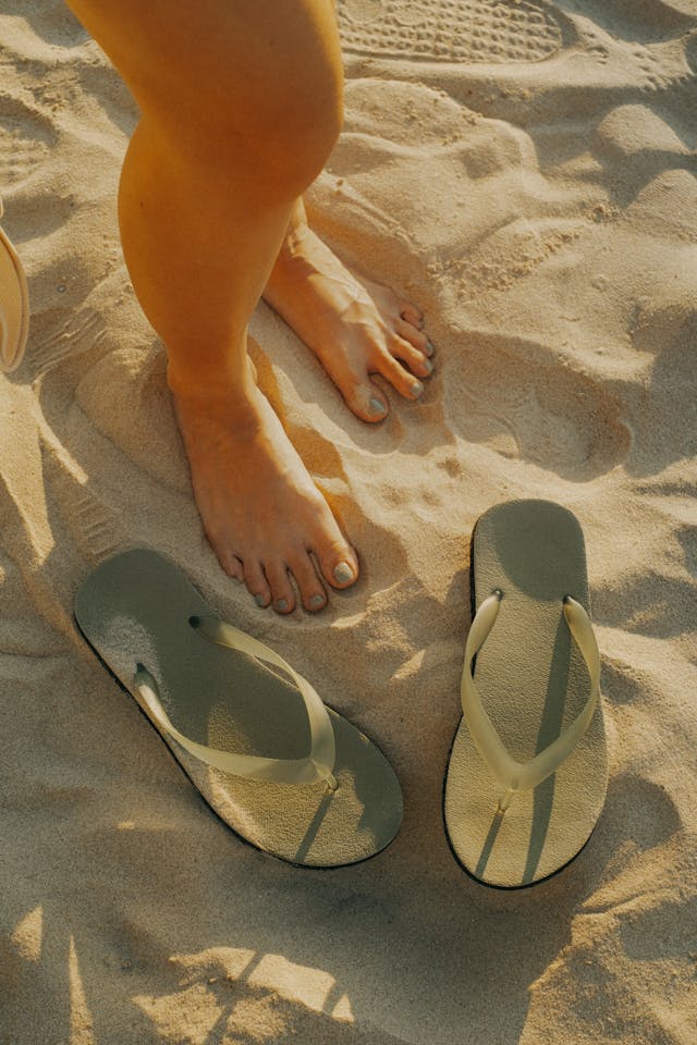 A pair of white and grey flip-flops lying in the sand next to the wearer on a beach.