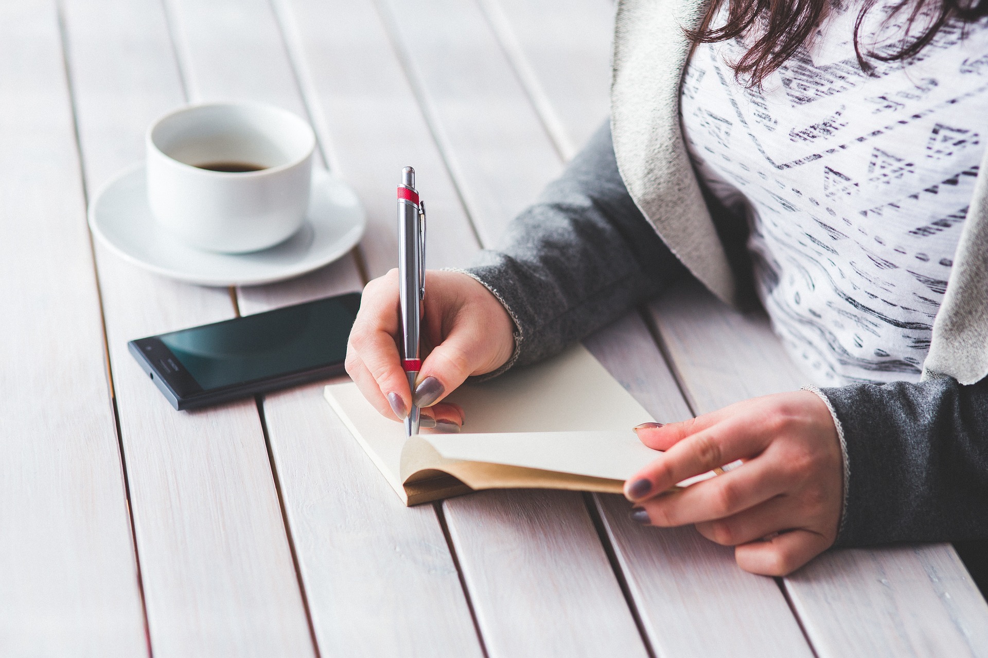 Female writing in a notepad with a mobile phone and cup of coffee on a table she is sat at