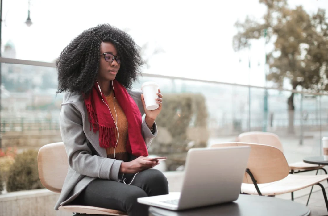 A woman drinking coffee and holding a phone sitting outside at a table looking at a Petzyo discount on the laptop