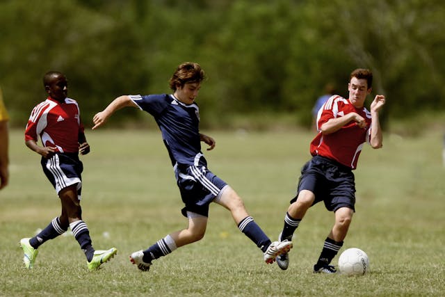 One man wearing a blue soccer jersey and two wearing red soccer jerseys playing soccer on a soccer field.