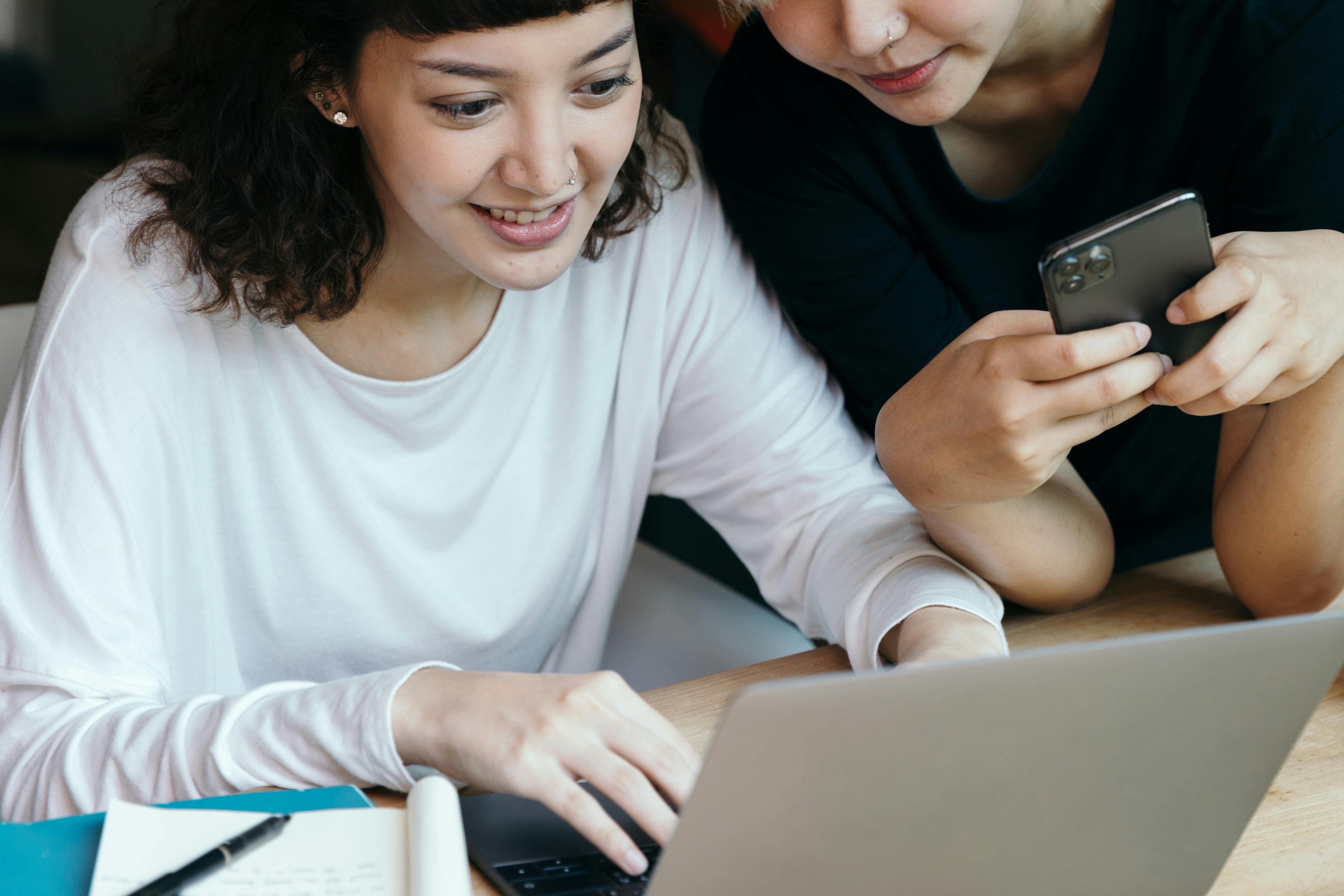 Two ladies sitting down, one typing on a laptop and the other holding a phone with hands.