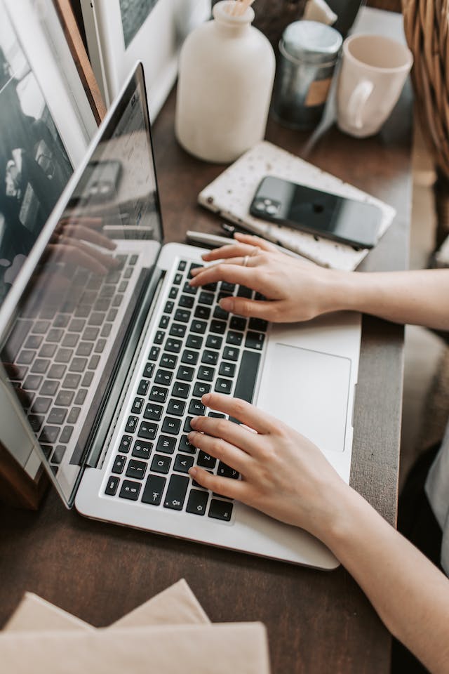 Person typing on a laptop while looking at a Whittard of Chelsea promo code with a mobile phone, cup and some paper with notes written on sitting on a wooden desk