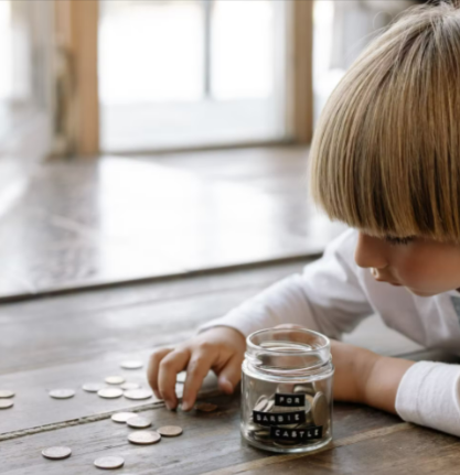 A young boy counting coins and placing them into a jar while sitting on wooden flooring.