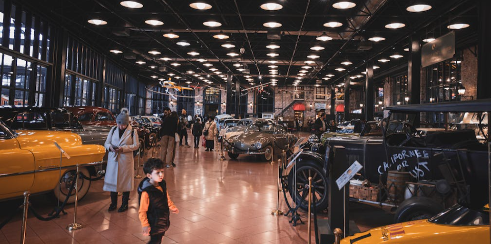 People in car showroom, vintage cars on display, bright ceiling lights. Cars for sale text is seen