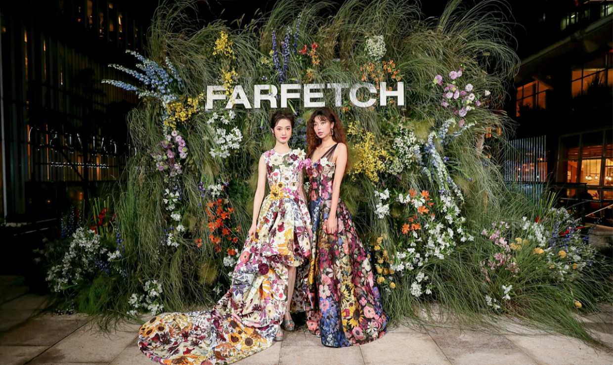 FARFETCH logo yazısının yanında çiçekli abiye elbise modelleri giyen iki kadın fotoğraf çektiriyor