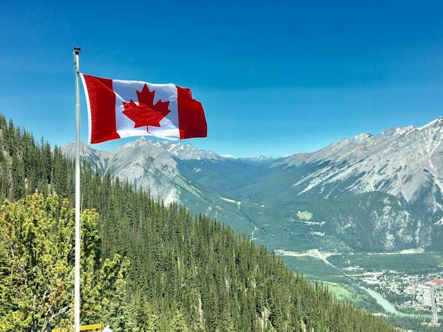 The red and white Canadian flag flying in the wind above a mountainous and forest landscape.