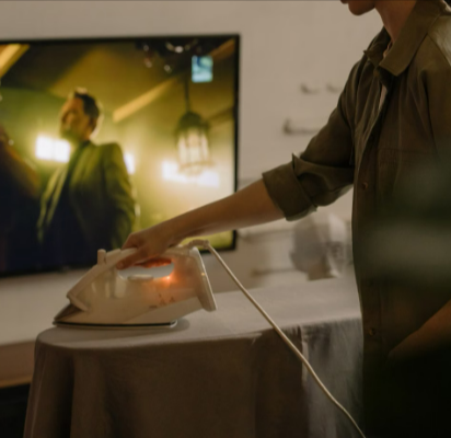 A woman irons clothes indoors while watching a television show with appliances bought from the Go Electrical savings section.