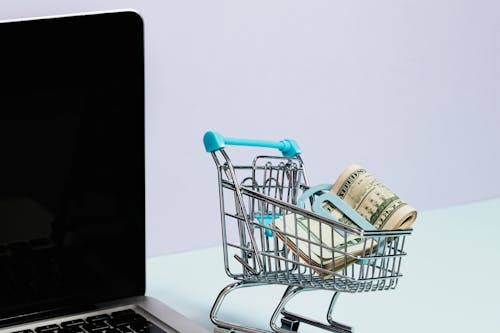 A small shopping cart filled with money next to a laptop.