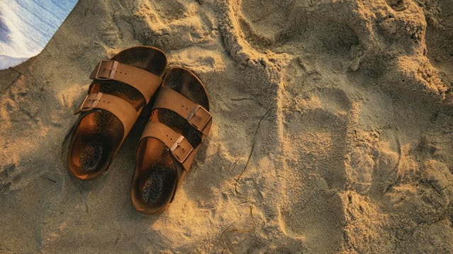 A pair of brown footbed sandals lying in the sand next to a beach towel on a beach.