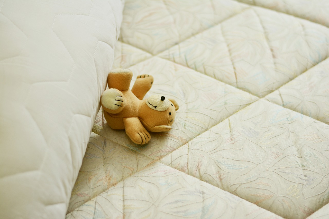 osito pequeño de peluche marrón sobre colchón color crema y blanco en posición dado vuelta de frente