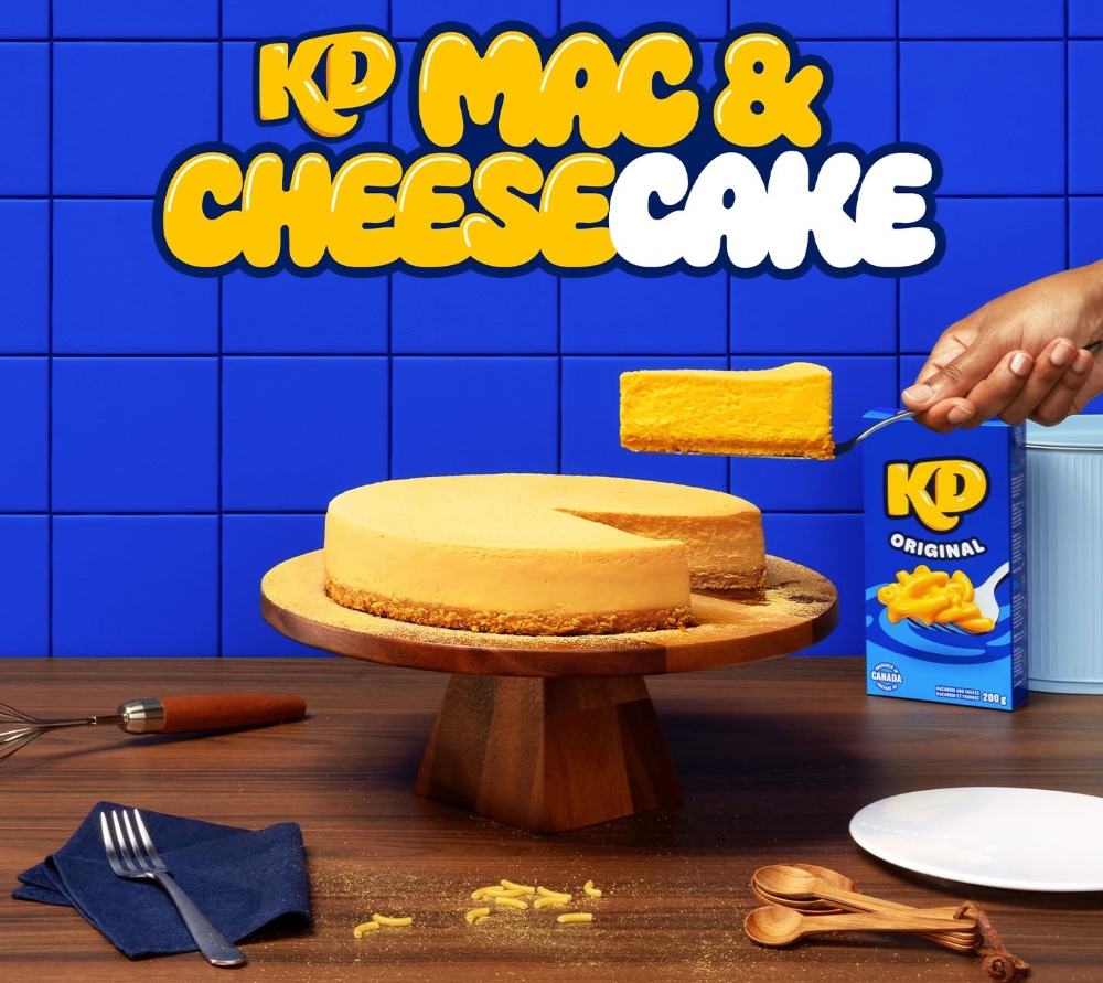 A KD Mac & Cheesecake resting on a cake stand with a slice being cut out of it