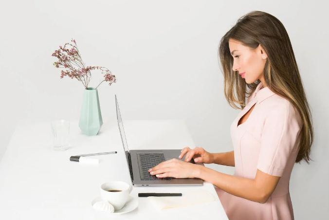 Lady with long hair in a pink dress using her laptop to search for the best Sculpted By Aimee Connolly voucher code