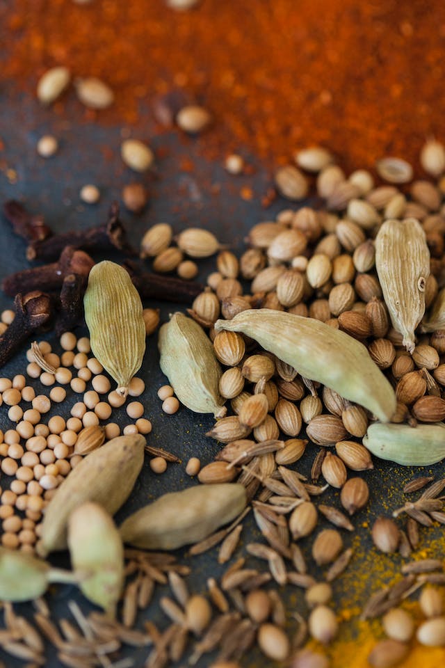 A pile of various types of seeds, seed pods, legumes, and spices lying on a dark stained table in a kitchen.