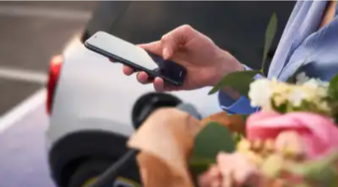 Eine Person neben einem Auto, die auf Ihrem Smartphone tippt und dabei Blumen hält