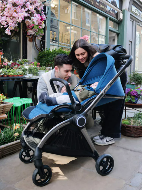 Parents playing with their baby in the street beside a cafe while the baby is in an iCandy blue and black pushchair from the bundle