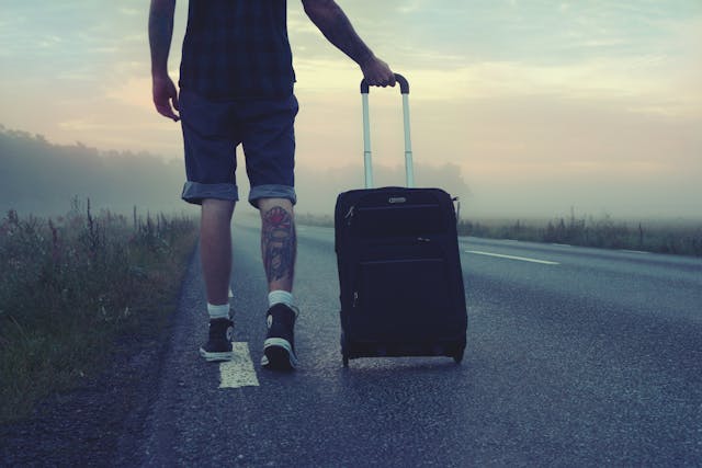A man with a tattoo on the back of his leg walking down a road with his black suitcase wheeling behind him