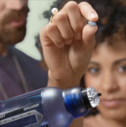 A woman wearing a ring holding a Bathmate hydropump and a blue pill with a man in the background