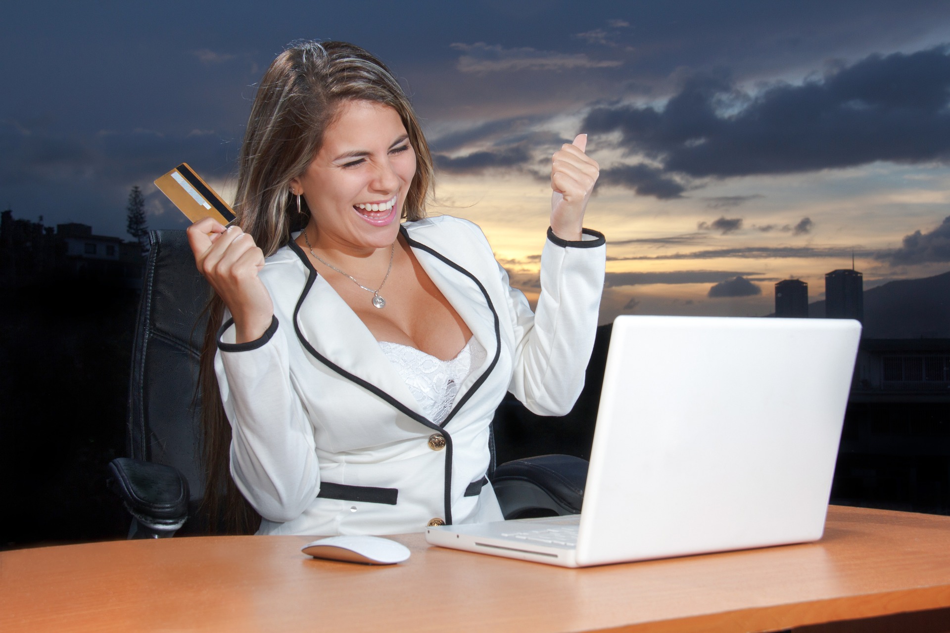 a woman wearing a white blazer looking excited while looking at a laptop with one of her hands holding a credit card