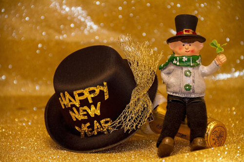 Eine kleine Figur mit Kleeblatt neben einem größeren Neujahr-Hut vor einem gold-glänzendem Hintergrund