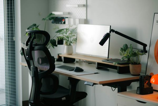 Black ergonomic mesh office chair in front of a desk with a monitor, keyboard, mouse, desk lamp, and a plant