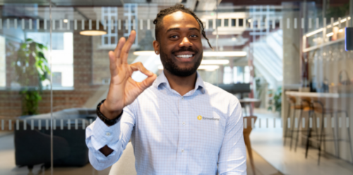 A man in an office smiling and giving an "okay" gesture with his right hand