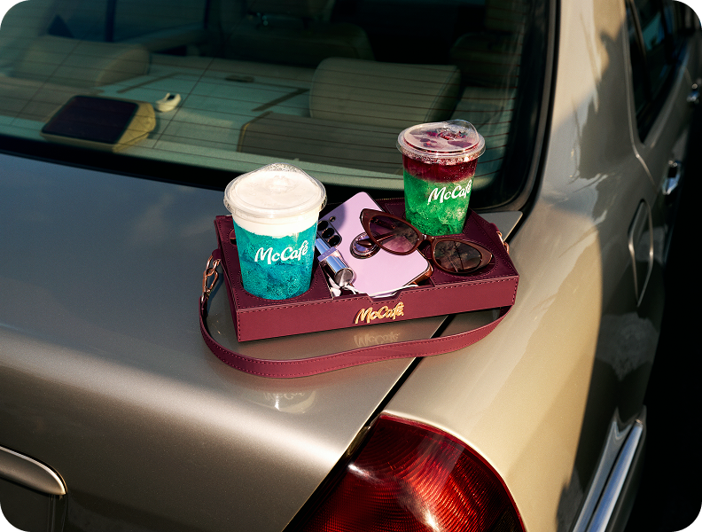 A McCafe tray bag resting on the trunk of a car and holding two drinks, a phone and sunglasses