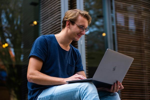 A white man sitting down holding an open laptop in his hands and smiling