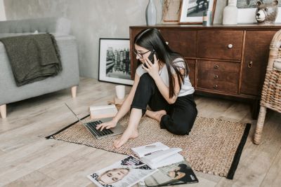 A young lady sitting on the floor with an open laptop, browsing on it while holding a phone in her ear