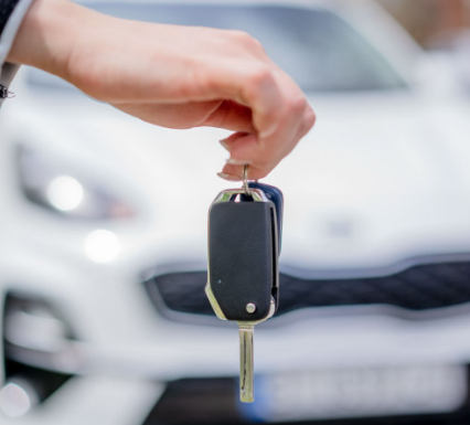 Close-up of a person holding a car key, with a white car in the background outdoors at an airport pickup.