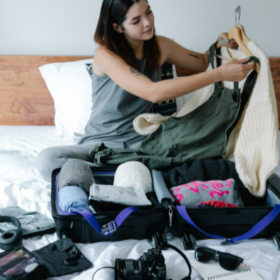 Young woman packing suitcase on bed, preparing for a travel adventure with clothes and camera equipment.